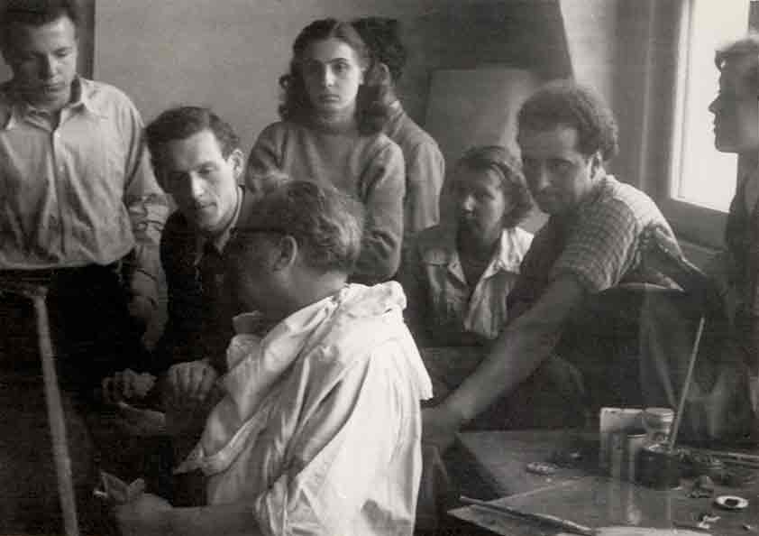 Foto: Abgebildet sind Gerhard Uhlig (zweiter von links) im Gespräch mit dem vor der Staffelei sitzenden Willi Baumeister (dritter von links) in der Staatlichen Akademie der Bildenden Künste Stuttgart im Jahr 1950 sowie weitere damalige Studenten. Copyright.